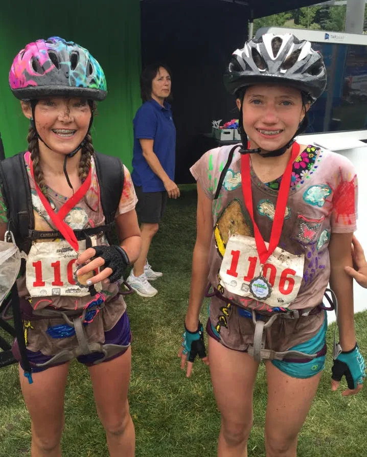 Two young racers in helmets, bib 1106, and finisher medals, caked in mud after the Kids Adventure Race in Vail